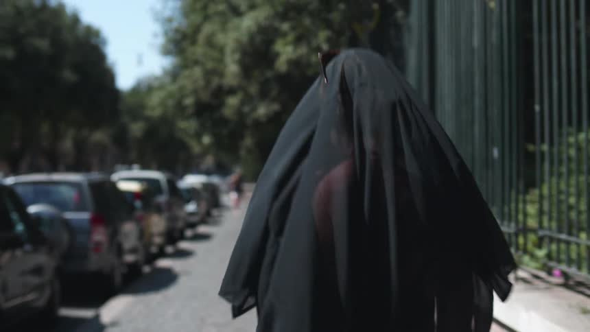 A woman wearing a lack scarf over her head walks along a sidewalk lined with vehicles