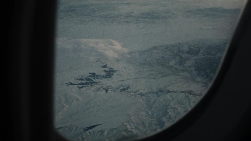 A camera is pointed at the window of a airplane as it flies over snow covered mountains