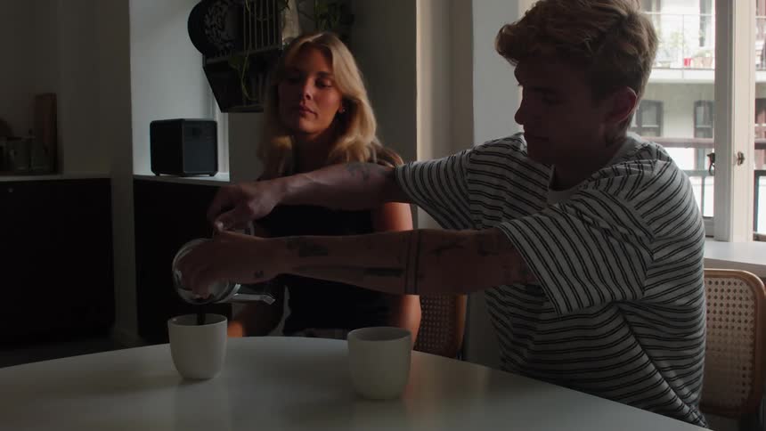 A young man pours coffee into a mug when a woman sits down beside him and smiles