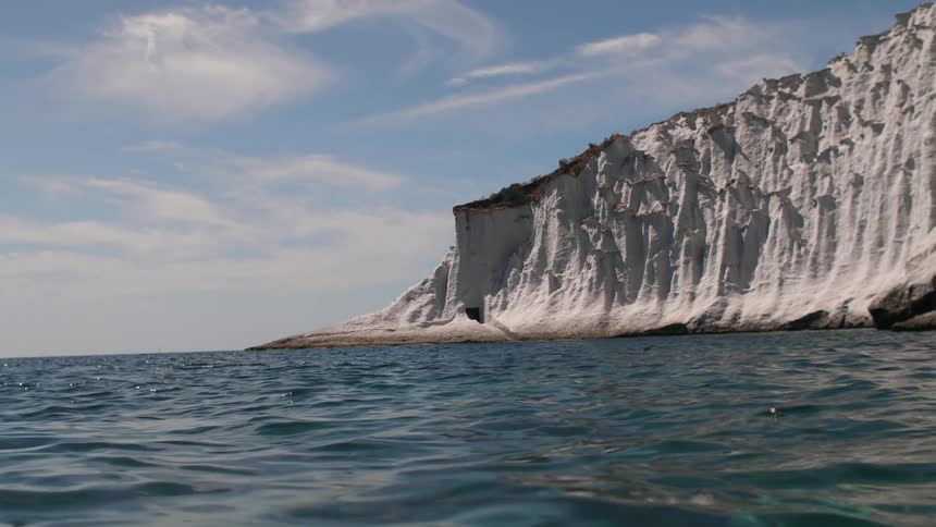 Steep oceanside cliffs contain a hidden cave under partly cloudy skies