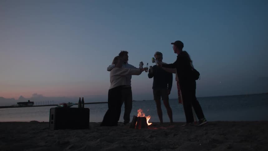 Five people drink on a beach next to a small campfire