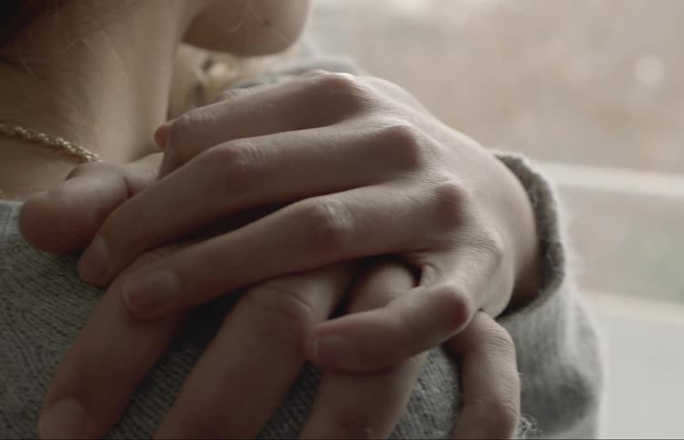 A man hugs a woman from behind as she looks out a window and she grabs his hand tight
