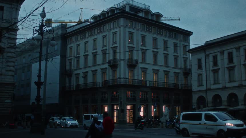 A large white building on a street with a few cars and people passing by in a twilight