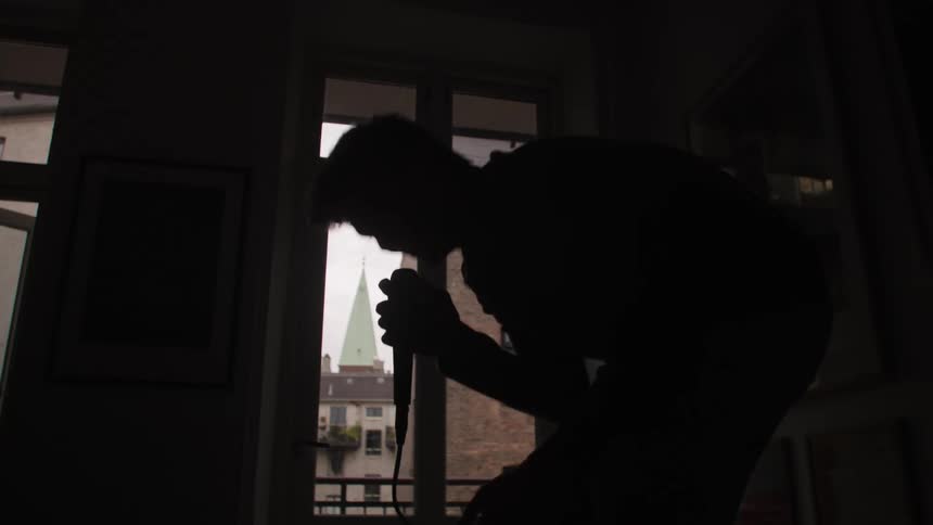 A man in silhouette in an apartment framed by light from a window bent over and singing into a microphone