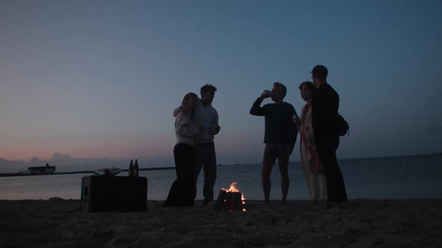 Group of friends standing on the beach sand drinking in front of a bonfire in the late afternoon sky