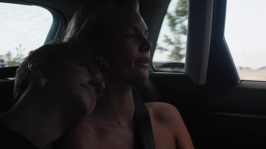 A couple sits chatting in a car as it drives down a highway the man rests his head on the womans shoulder