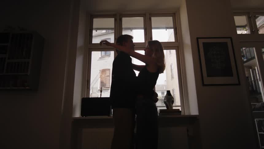 A woman approaches a man who is leaning against the window and they hug and look at each other romantically