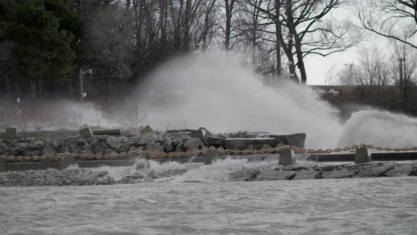 Birds are flying and hovering as waves crash across a jetty on a cloudy day