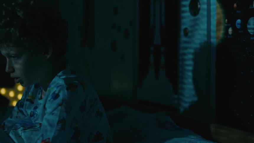 Boy in pajamas sits up on bed and sees a shadow creping across the wall opposite his window