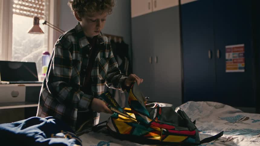 A male child arranging school items inside his backpack in the bedroom at midday