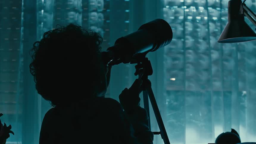 A silhouette of a boy using a telescope inside a room