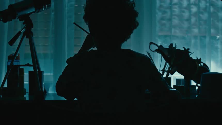 Boy sitting at desk is silhouetted against the windows in dark room