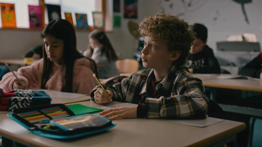 A boy is writing with a pencil while sitting in a classroom