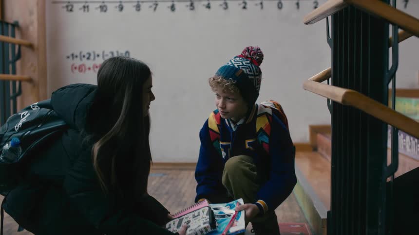A boy with a backpack helps a girl gather several books that are on the floor and then he goes up the stairs