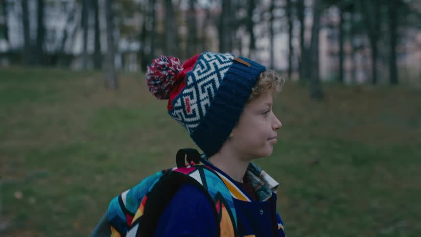 A boy in a colorful jacket and beanie walks near a forest