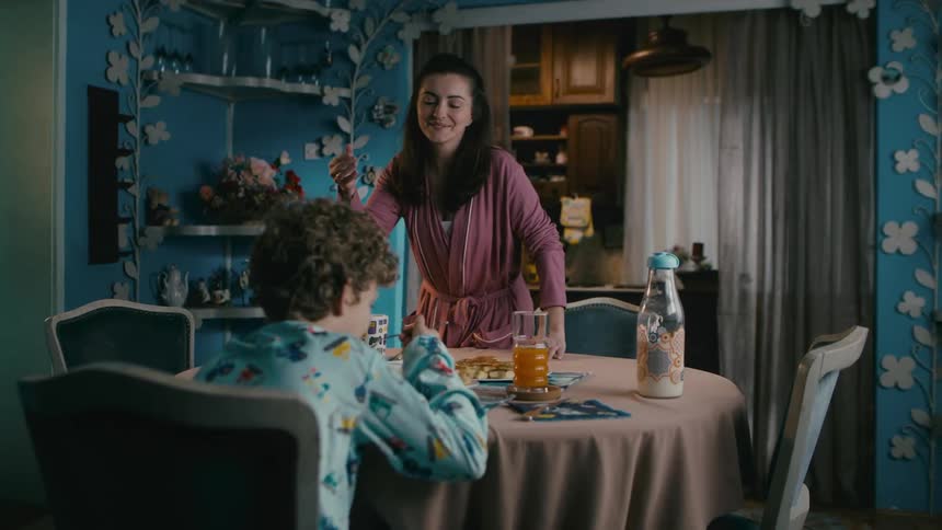 A mother brings food to her son at the kitchen table