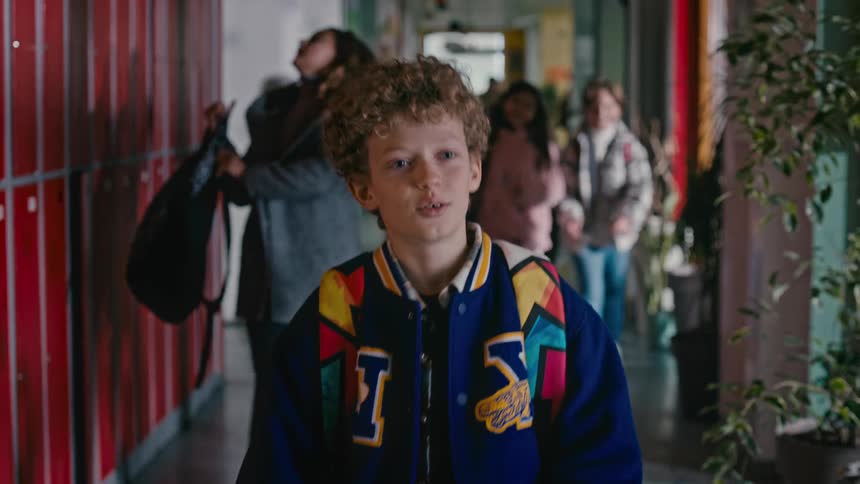 A young boy with curly hair walks through the hallway at school and waves to other kids