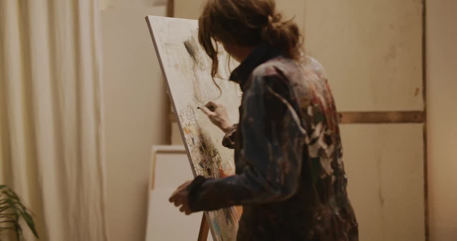 Female artist stands and draws on canvas board set on easel in curtained room