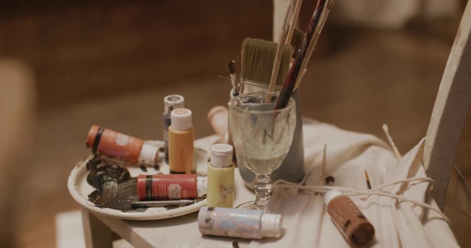 Paints and brushes spread on a small table near an easel with a canvas
