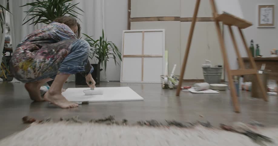 Woman crouched on the floor applying a roller of white paint to a canvas