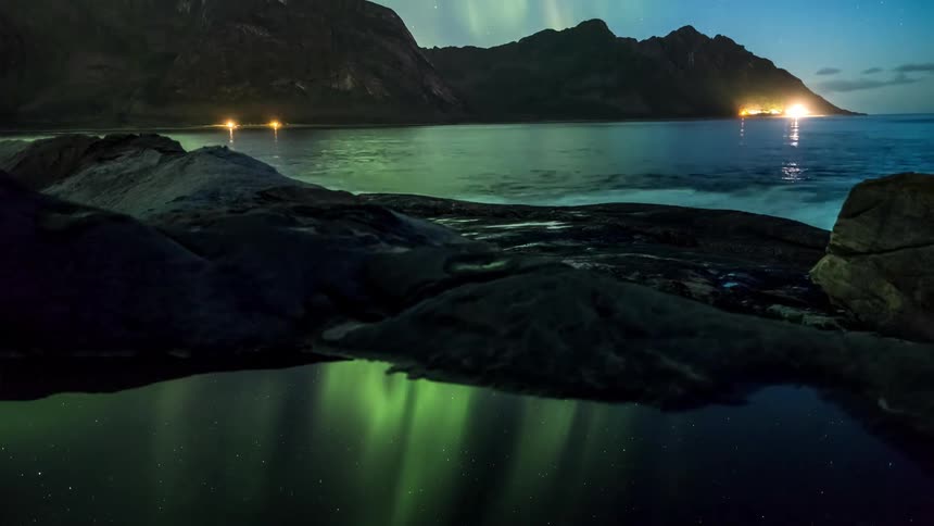 The lights of an aurora borealis reflecting in the waters of a lake between mountains