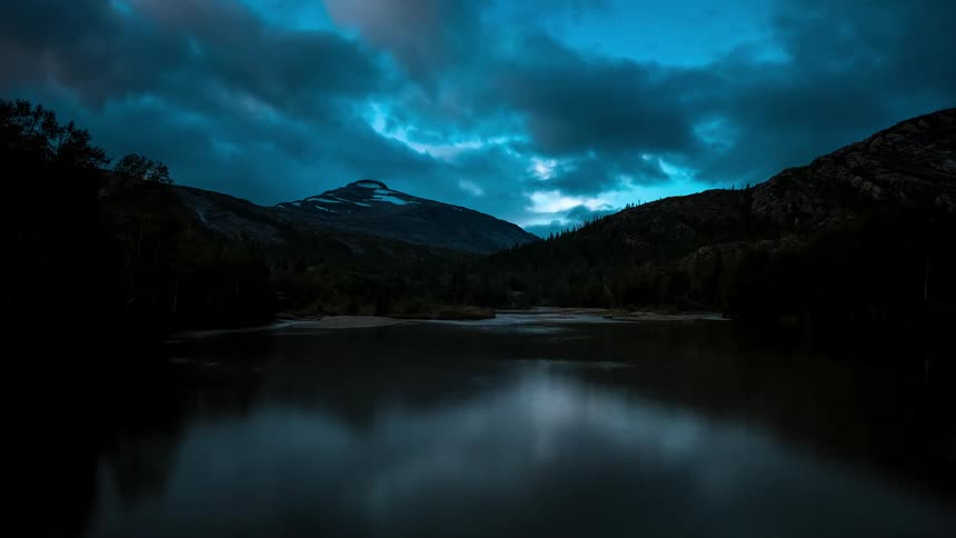 Landscape of a lake with forests and mountains around it and the sky darkening and dawning in timelapse