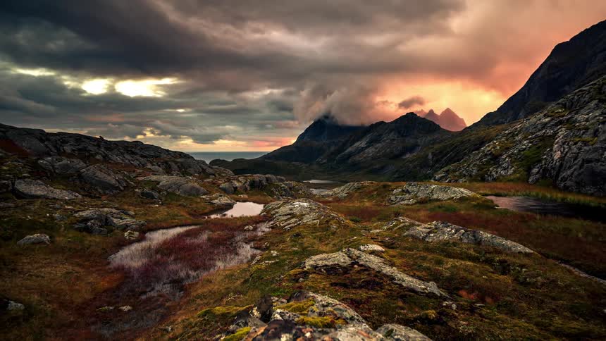 A region with the ground covered in vegetation and rocks and behind mountains and many clouds passing in the sky