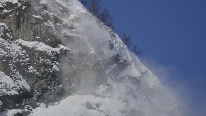 Mountain with cliff with snow flying through the air on a cloudless blue sky day