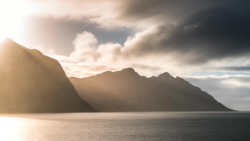 Two huge mountains facing the sea and one receiving sunlight while several gray clouds pass in the sky