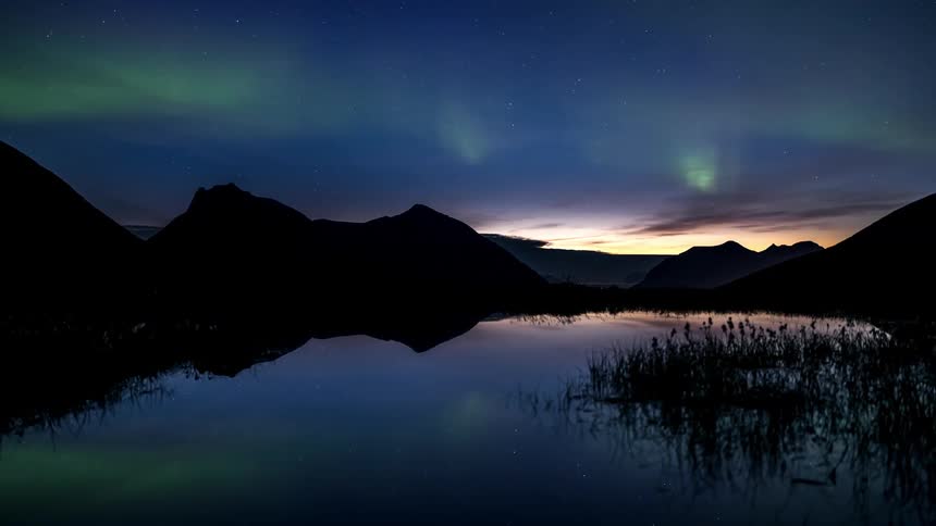 Northern lights reflected in the water of the lake surrounded by mountains during the starry night