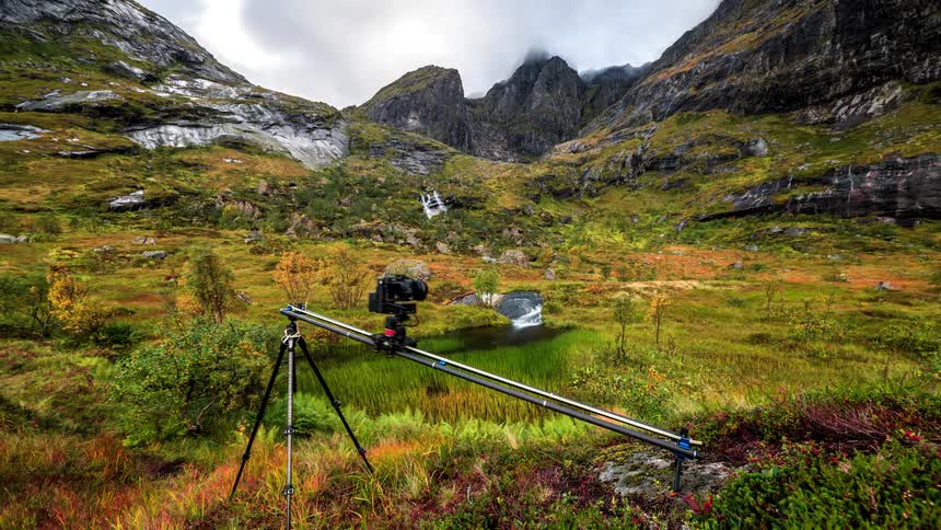 Man wearing a blue jacket and black pants puts camera on tripod to film the mountainous area on a cloudy day