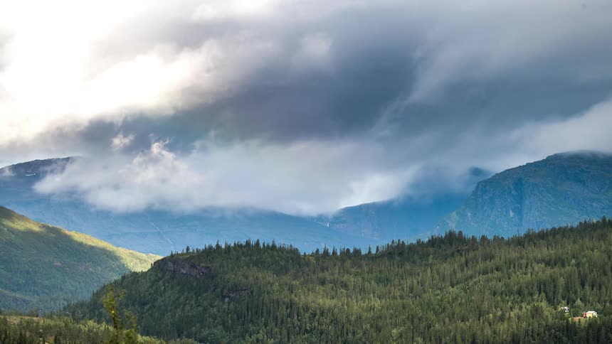 A mountainous region with some forests of trees and a dense cloud in the sky going to the right