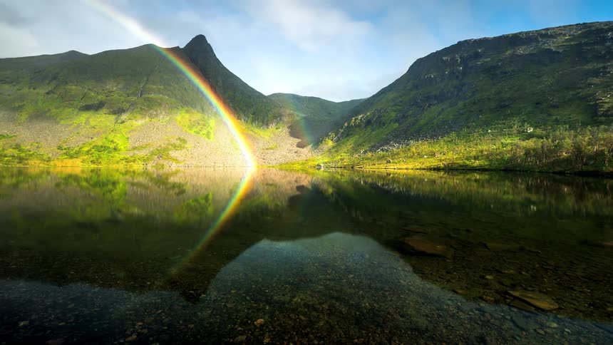 Landscape of a reflective lake with rainbows and mountains with forest