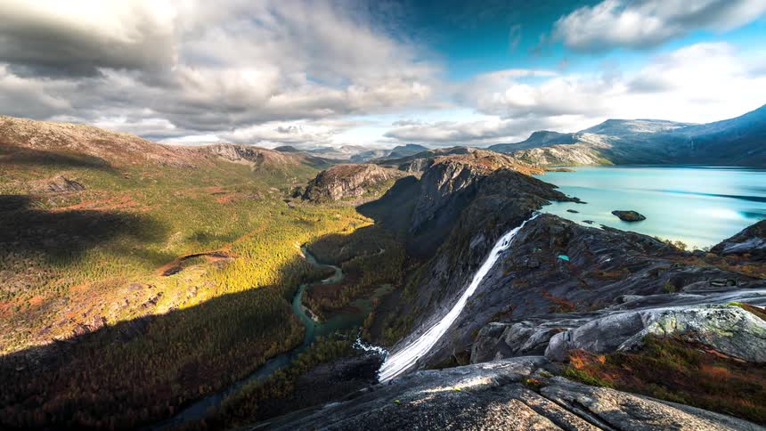 Landscape of mountain ranges with vegetation with a turquoise lake giving access to a waterfall and clouds passing by in timelapse