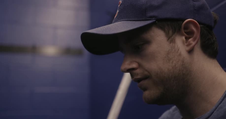 A man sits in a locker room wearing a baseball cap