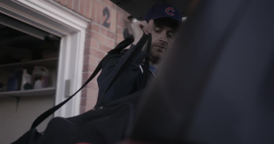 A man wearing a chicago cubs baseball cup loads a bag into the trunk of a car
