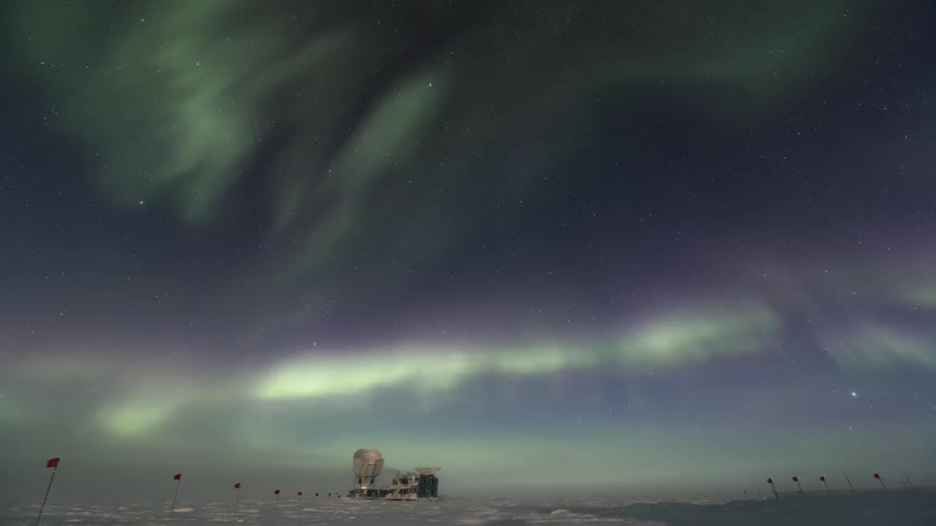 The Aurora Australis with a green glow moving in the sky over a region with snow on the ground and some flags