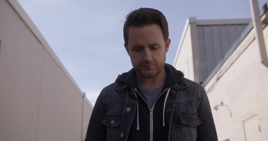 A man wearing a denim jacket walks down an alley looking sad