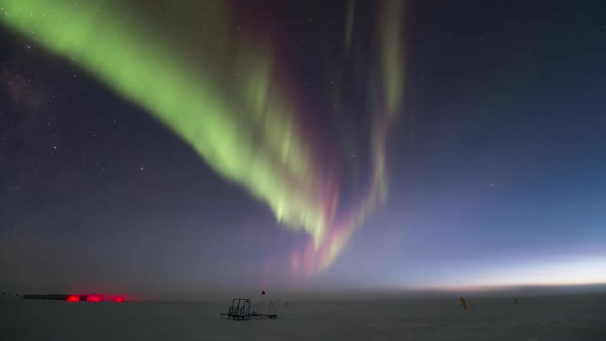 Aurora Australis over a research station and a snowy field marked with flags at night