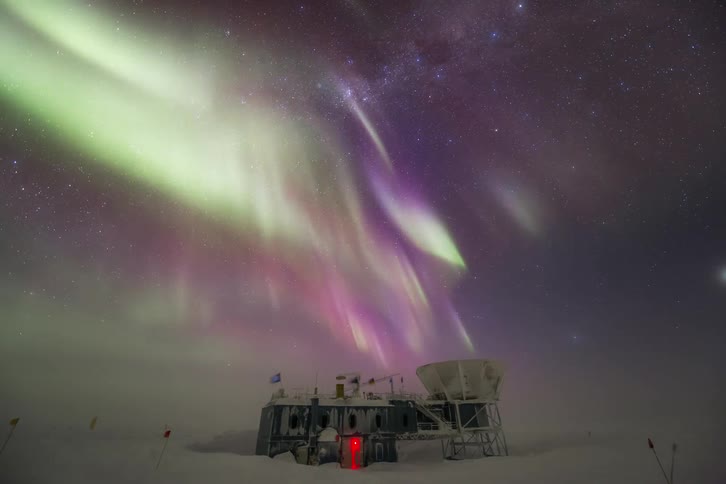 A bright  Aurora Australis in the sky near a building in the snow with a person observing and placing a camera on a tripod to record