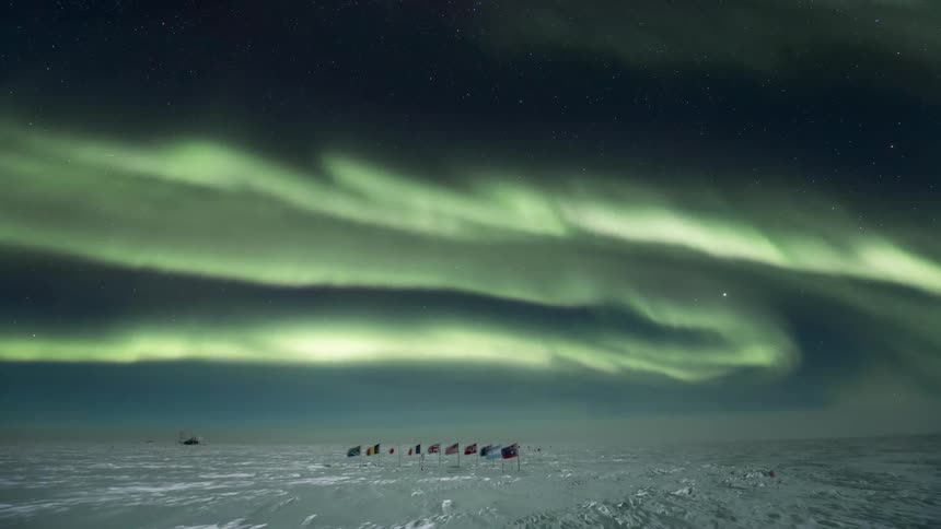 An aurora swirls in the sky over a row of flags planted in the snow