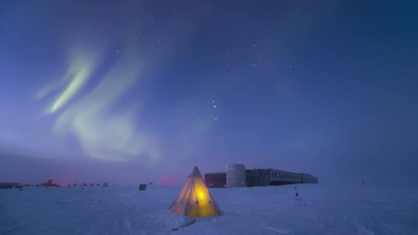 Aurora Australis shining over a research station and a snow field with a tent