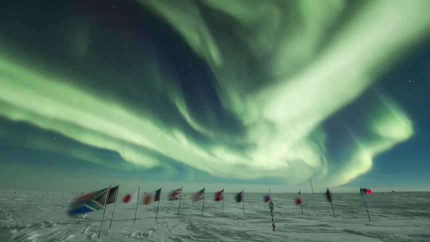 Aurora Australis wave in blue sky above a row of various nations flags planted in snow covered ground