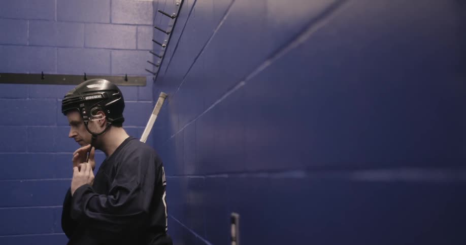 A man sitting in a locker room is putting on a hockey helmet