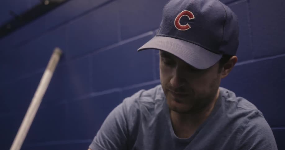 A man wearing a chicago cubs baseball cup sits on a bench in a locker room