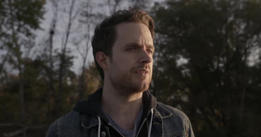 A man is standing in a wooded area staring into the distance