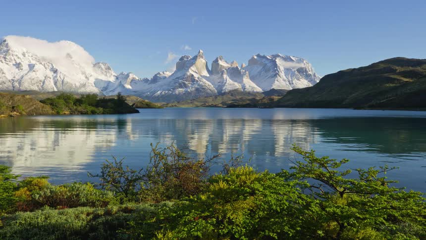 A landscape featuring a clear lake reflecting majestic snowcapped mountains and a blue sky
