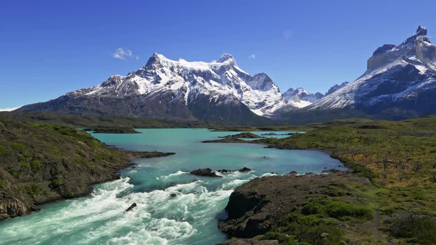 A river with rapids running through a grassy plain with a mountain range in the distance