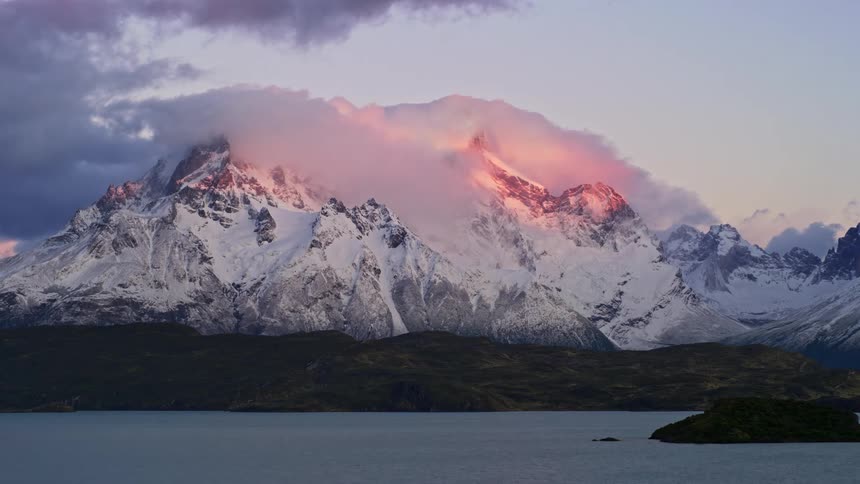 Timelapse of sun rising on clouds over a snow covered mountain beyond a lakeshore