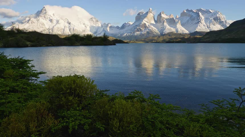 Green vegetation on the shoreline of a clear rippling lake reflecting a clear blue sky and peaks of white mountains in the background
